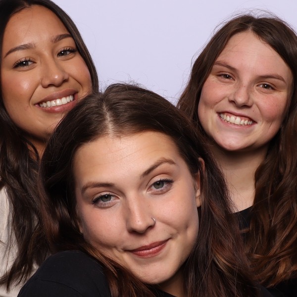 Close-up of three smiling women with long hair against a pale lavender backdrop