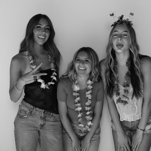 Black-and-white shot of three women in flower leis, two sticking out tongues, one in a Grad 2026 headband