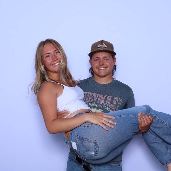 A guest in a brown cap carries a smiling woman in jeans against a plain light backdrop